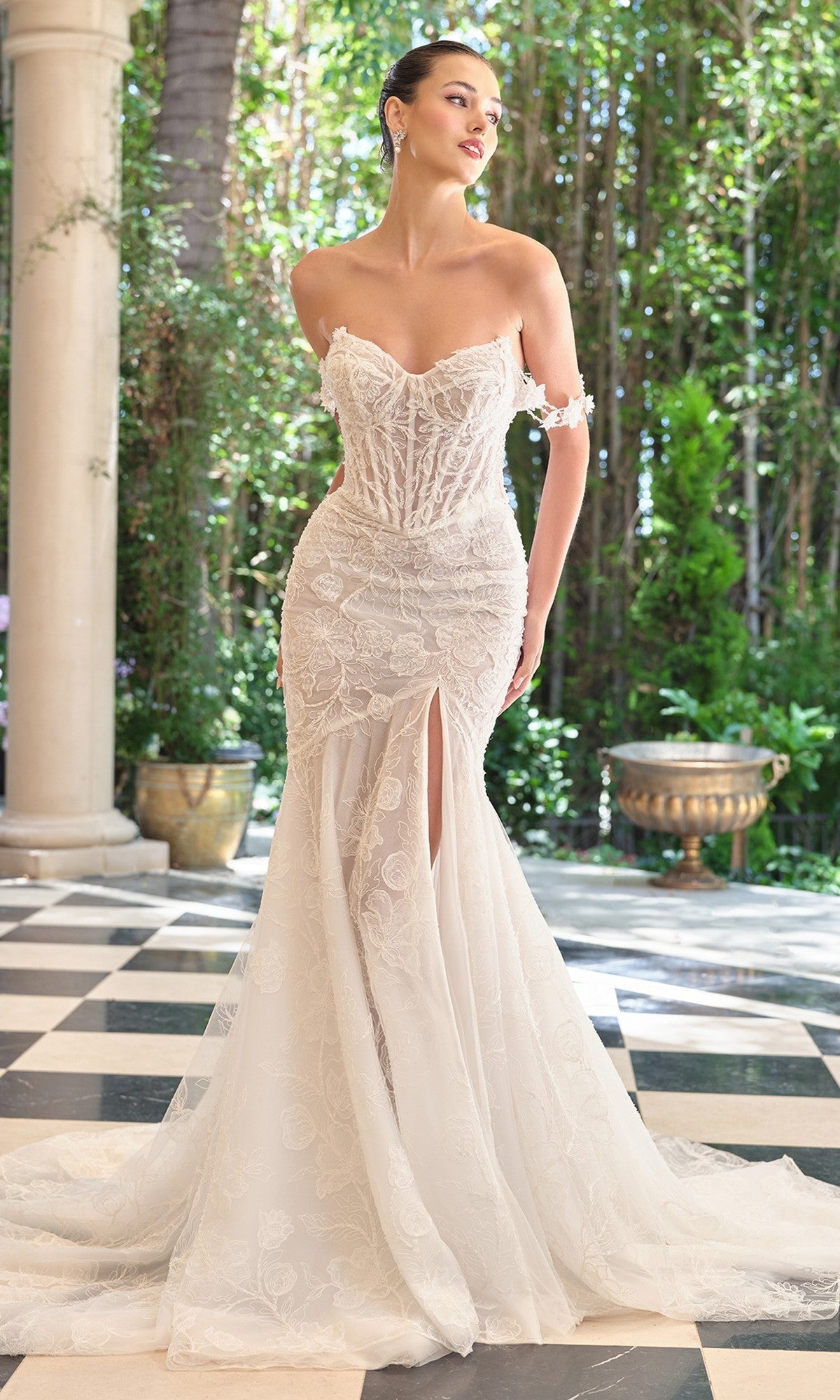 A woman stands on a checkered floor in a garden, wearing the off-white Andrea and Leo WL099 wedding dress featuring an off-the-shoulder neckline, floral lace details, mermaid silhouette, and a stylish front slit.